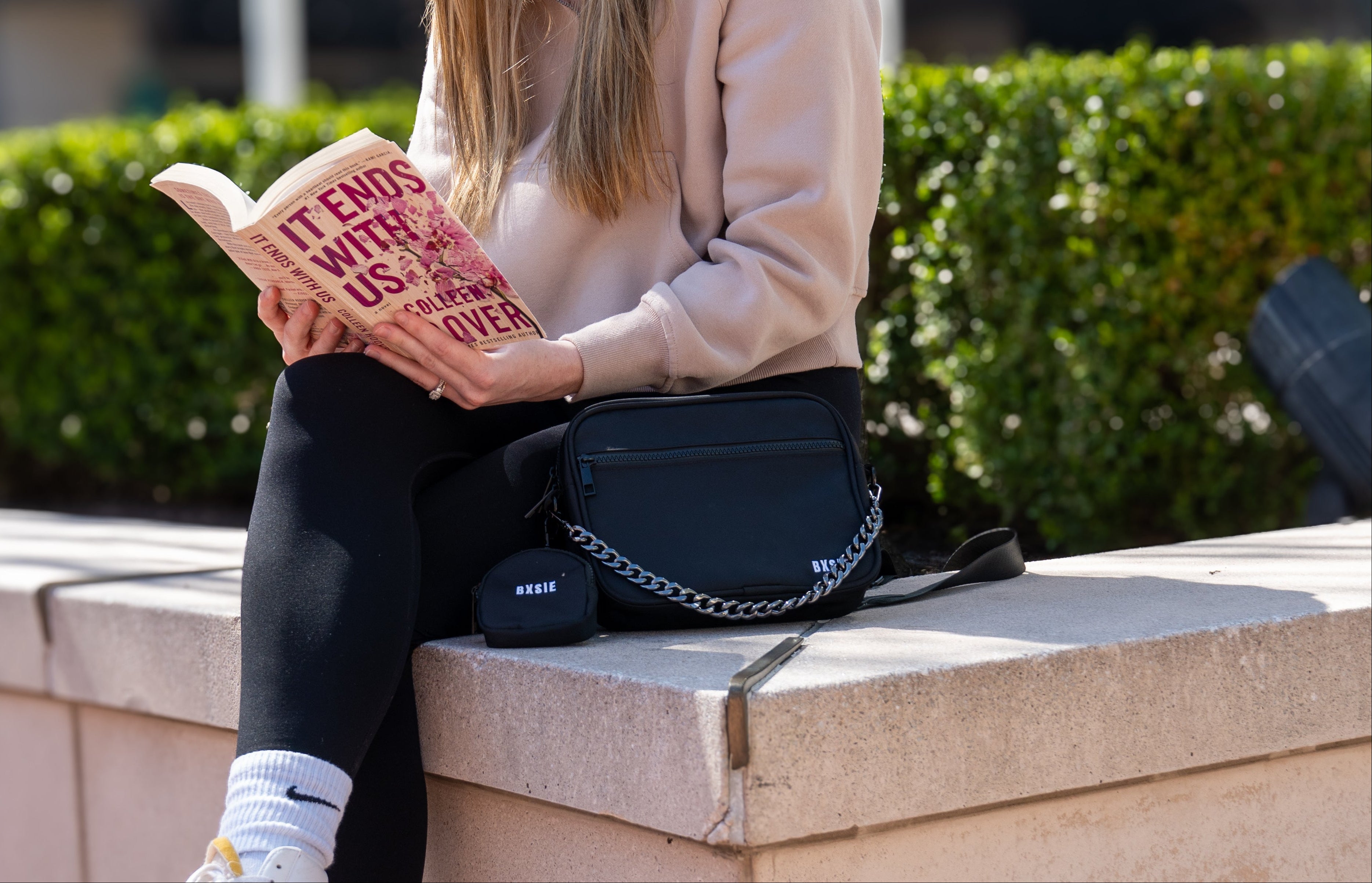 Woman reading a book outdoors on a concrete ledge with greenery and buildings in the background, with a BXSIE Kindle bag purse next to her.